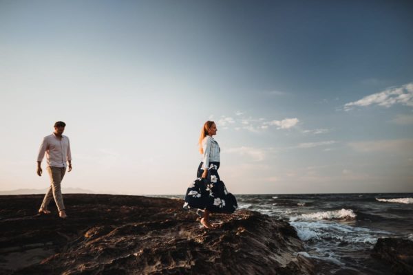 The bride is standing on brown rocks, looking out to sea and her groom is walking towards her.