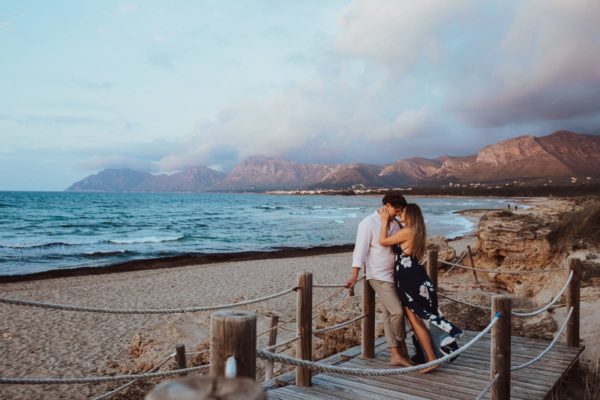 The bride and groom cuddle on the wooden walkway leading from the beach to the dunes. We see Majorca's wonderful blue sea and the mountains in the evening light in the background.