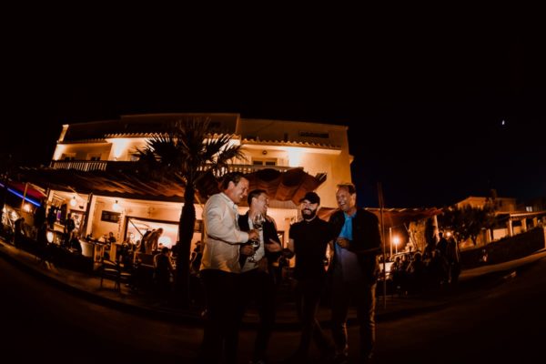 The brother of the groom and three friends laugh heartily in front of the illuminated Bar Sol in the fishing village of Son Serra de Marina in Majorca.