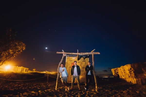 Group photo of the groom with his two best friends in the moonlight and blue hour in front of a server wooden house right on the beach.