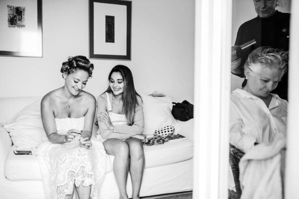 While the bride´s mum is going styled, the bride sits smiling on the bed with a bridesmaid.