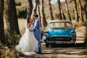 The hugging newlyweds in front of the classic car among green pine trees.