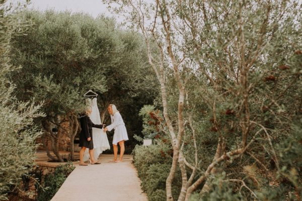 The bdrial dress is hanging on a olive tree. The bride and her mother are laughing.