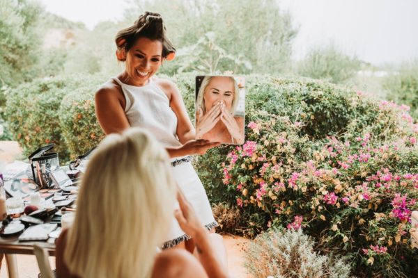 The make up artist lift up a mirror that the bride can see the result. She is smiling very happy.